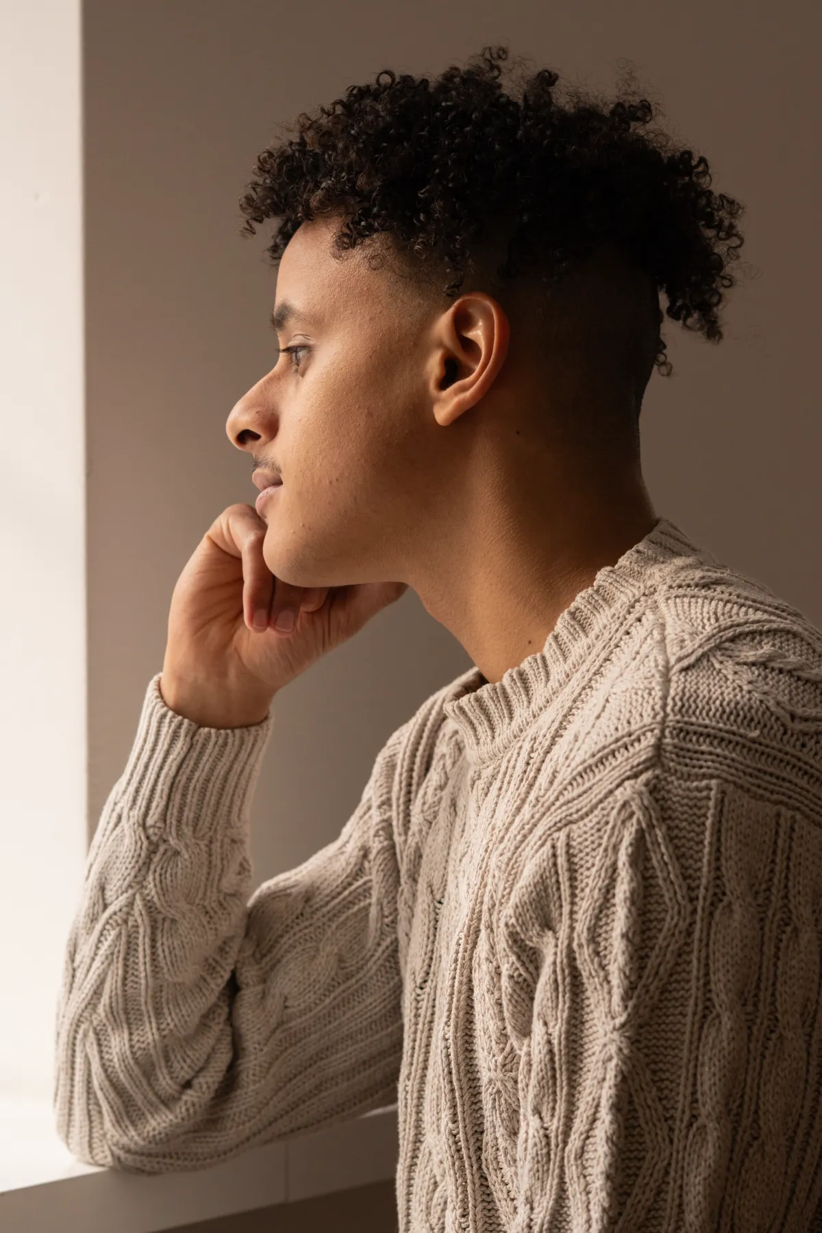 Profilporträt eines jungen Mannes im Strickpullover, der im weichen Fensterlicht nach draussen blickt